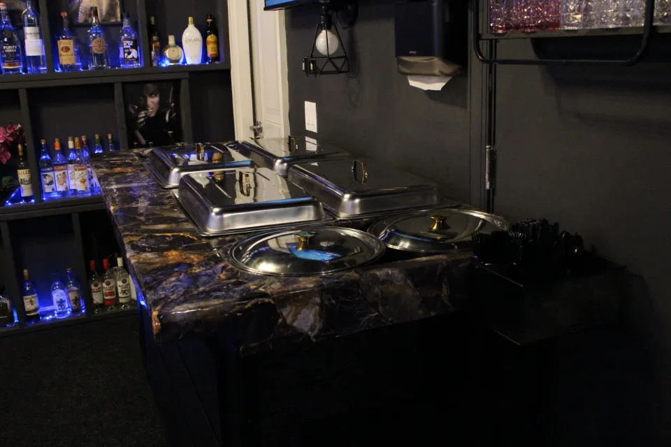 A buffet featuring hot dishes on a marble counter, with shelves of liquor bottles in the background.