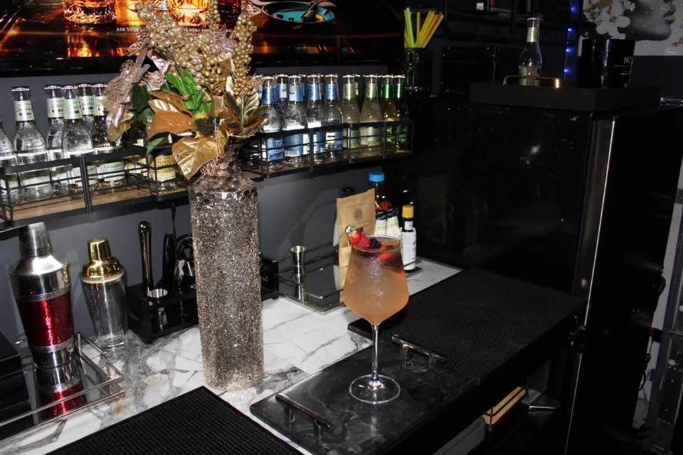 A cocktail glass with red berries and ice cubes, placed on a tray, along with a shaker and a floral arrangement on a bar.