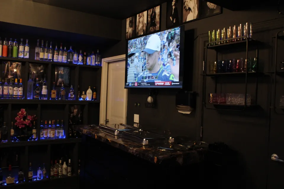 A bar area with shelves filled with illuminated bottles of liquor, a TV screen showing an American football game, and colorful glasses on metal shelves.