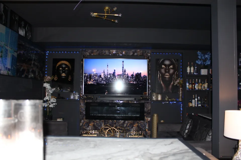A modern living room decorated with works of art, a well-stocked bar, and a fireplace flanking a television displaying a nighttime cityscape.