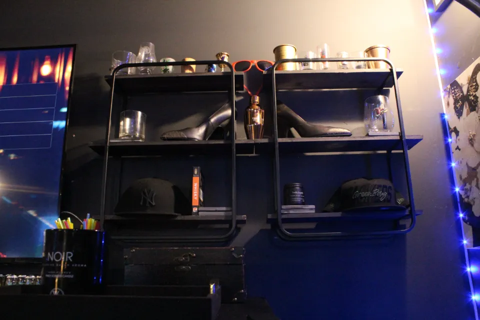 Black wall shelves displaying high-heeled shoes, caps, glasses, a bottle of perfume, and bar accessories.