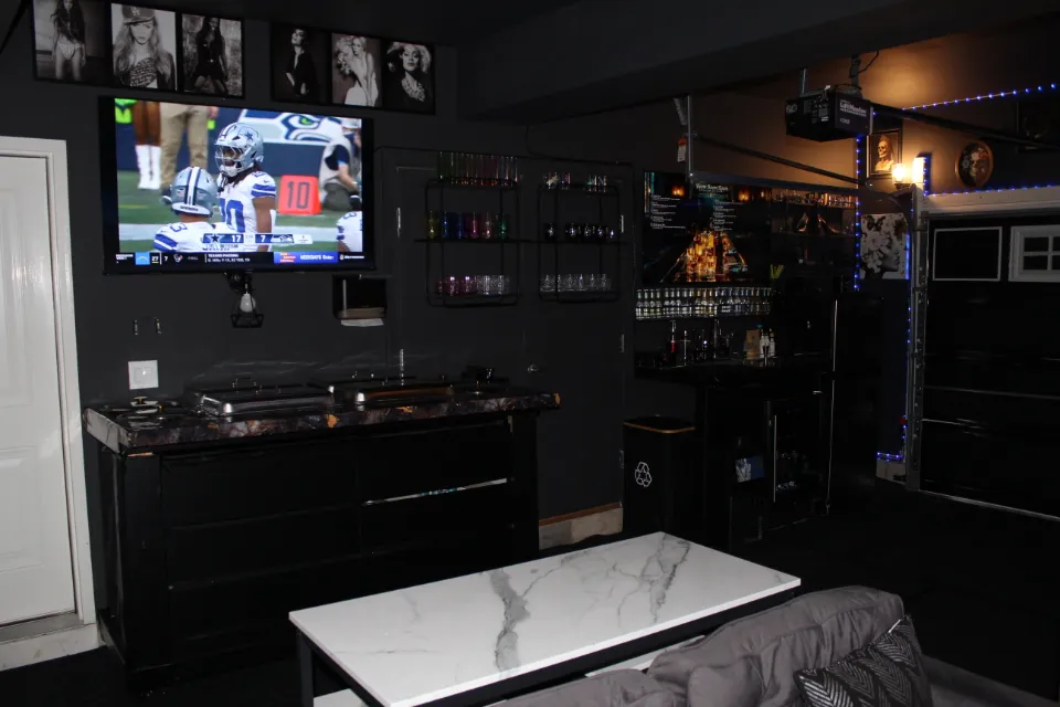 A dimly lit game room with a large TV screen showing a soccer game, a bar with shelves of drinks, and a marble coffee table.